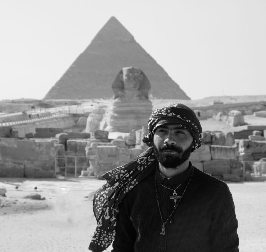 Meka from Meka Egypt Tours stands in front of the Sphinx and the Pyramids in a black and white image.